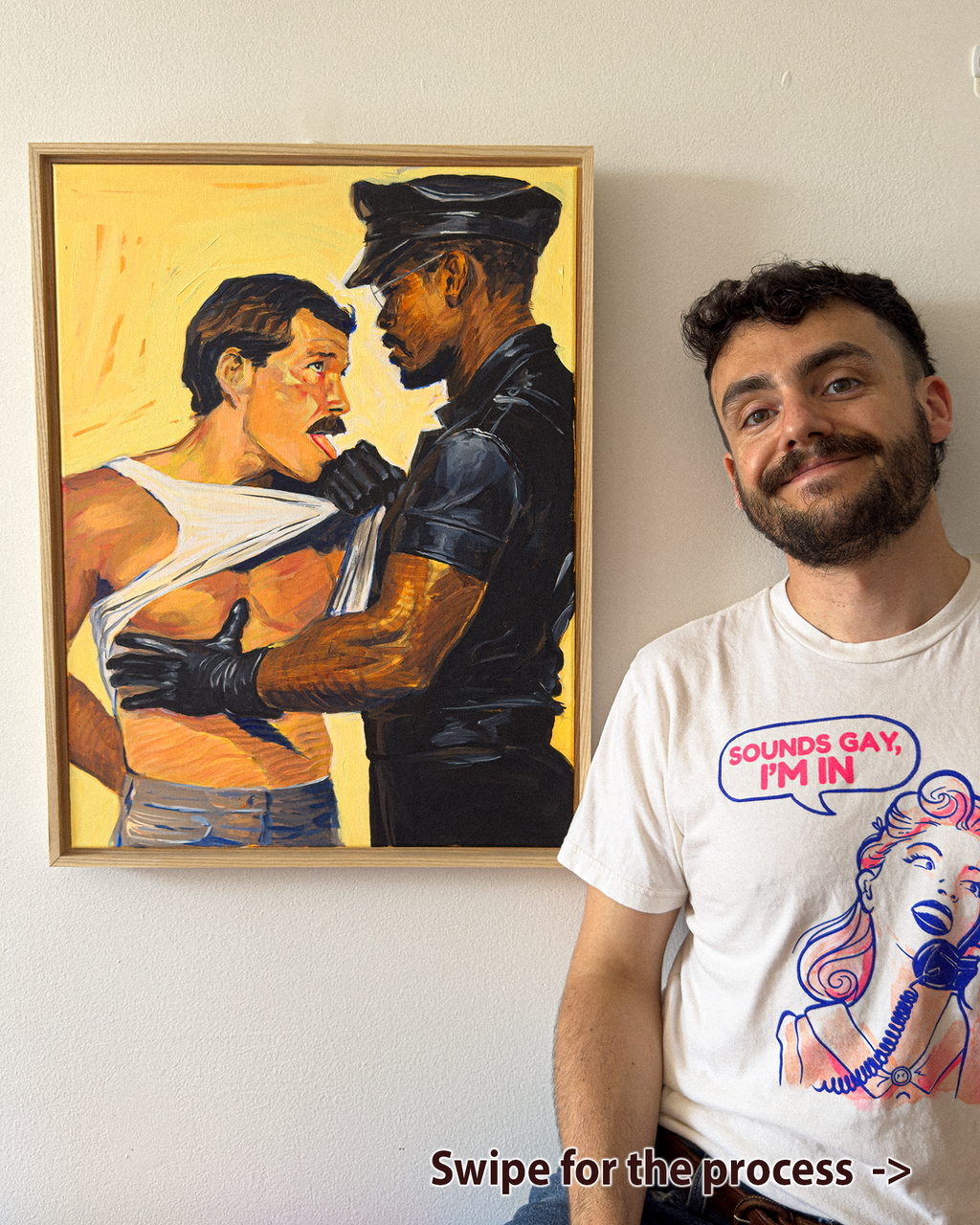 A man wearing a white graphic t-shirt stands next to a framed painting on a white wall. The painting depicts a man in a white tank top and a police officer in black.