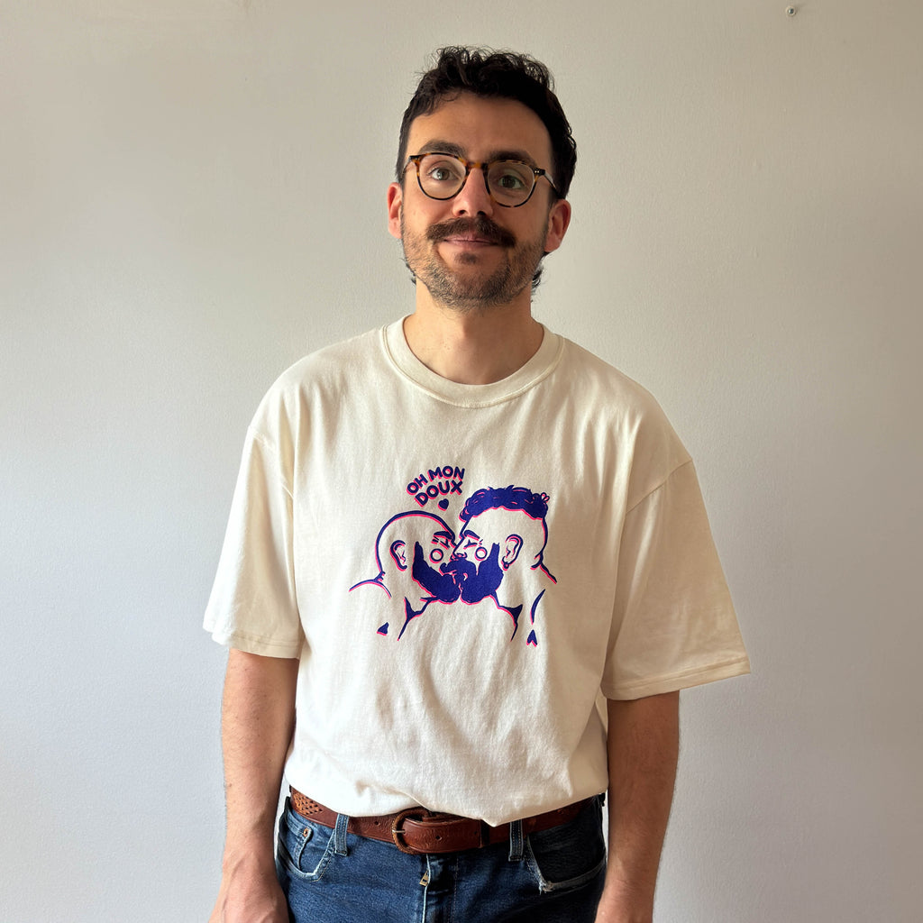 A man wearing a cream-colored graphic t-shirt with an illustration of two bear gay guys kissing cartoon and the text OH MON DOUX on the front, paired with blue jeans and glasses, standing against a plain light gray wall.