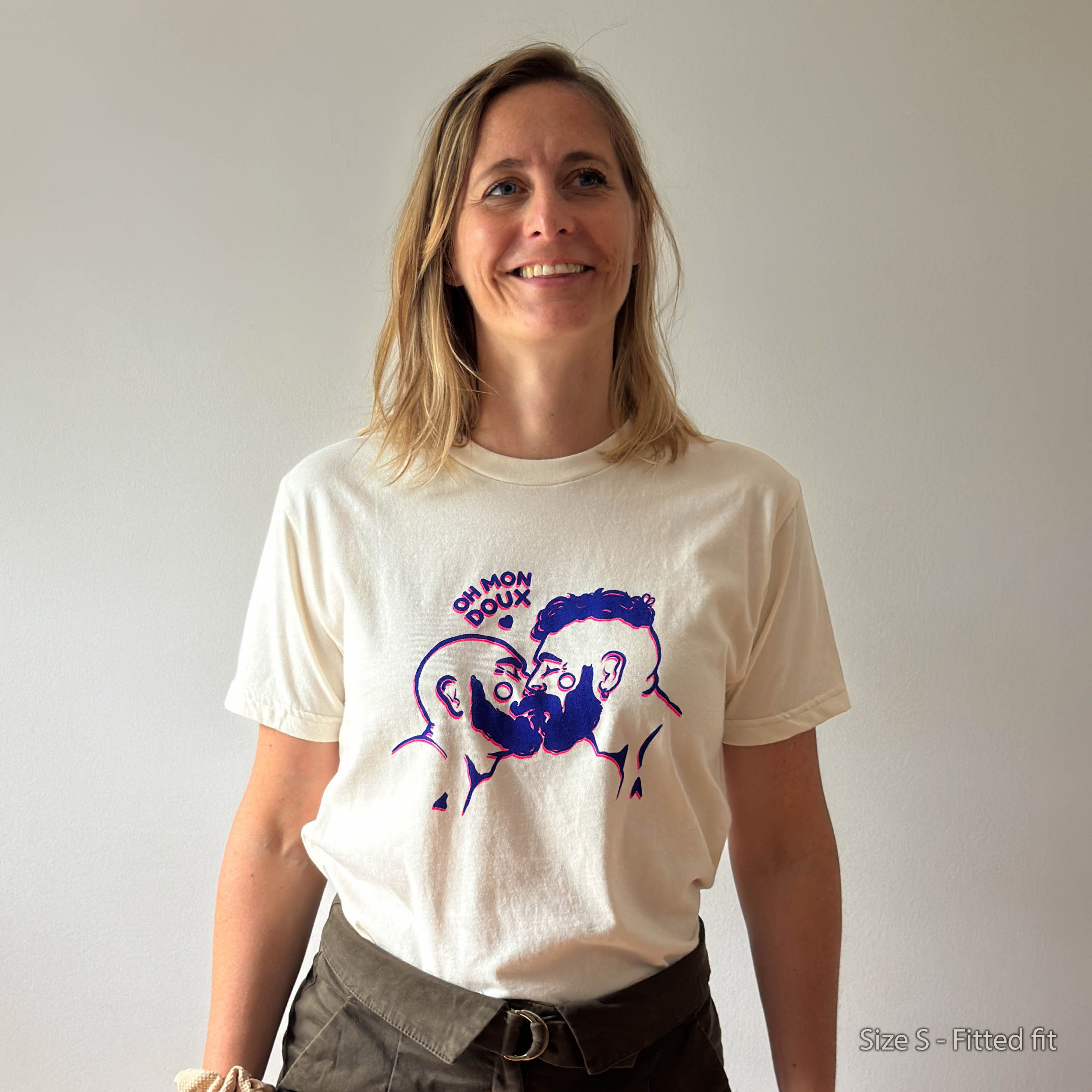 A woman wearing a cream-colored t-shirt with a purple graphic of two cartoon pigs and the text 'OH MON DOUX' on the front, paired with dark brown pants, standing against a plain light gray wall.