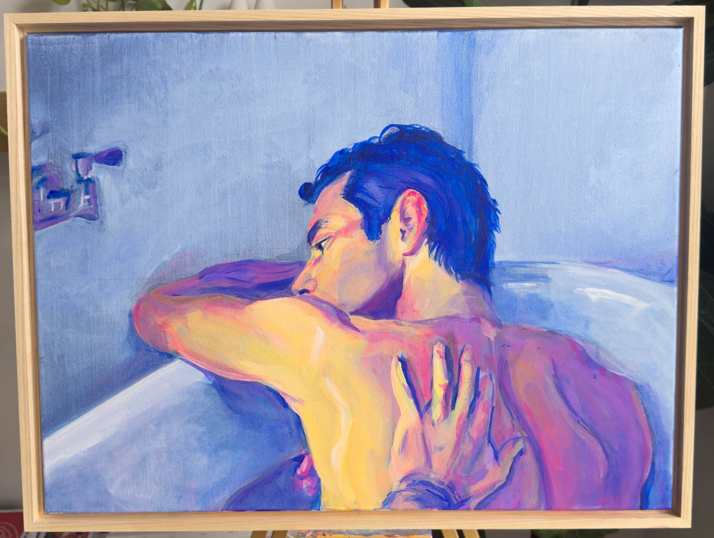 A framed painting on an easel depicting a man in a bathtub, rendered in expressive brushstrokes of blue, purple, pink, and yellow. The painting is housed in a light natural wood frame, set against a softly blurred indoor background with green plants visible.