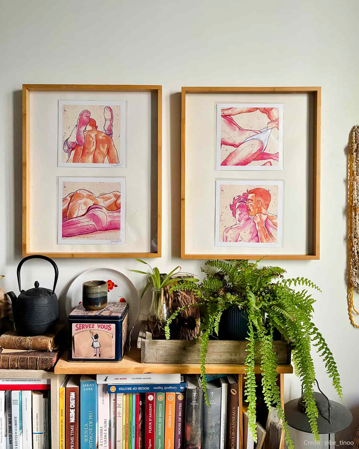 Four framed gay art prints depicting intimate moments between men, displayed above a bookshelf with books and houseplants.